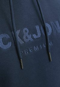 Sudadera navy con el logo bordado "CK&JON" en un hilo ligeramente más claro. Presenta cordones texturizados con acentos metálicos y una tela suave.