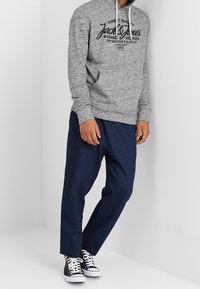 Sudadera gris con cordón, que presenta una impresión de logo negro. Combinada con pantalones oscuros de corte holgado en azul marino y zapatillas negras.