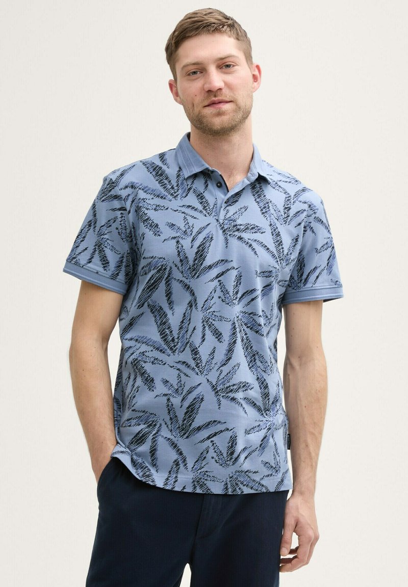 Polo bleu à manches courtes avec un motif de feuilles noires, col classique et tissu texturé. Porté par un mannequin avec les mains dans les poches.