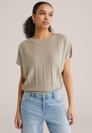 Donna con capelli ricci che indossa orecchini a cerchio d'argento, un top beige a maniche corte lavorato a maglia e jeans a vita alta azzurri, che guarda leggermente a destra.