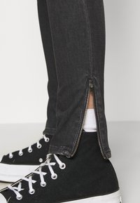 Vaqueros de mezclilla negros con cremalleras en los laterales a la altura de los tobillos. Combinados con zapatillas altas negras y calcetines blancos. Diseño clásico ajustado.