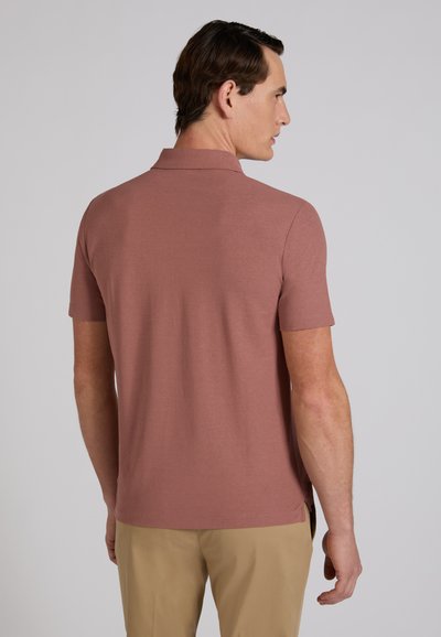 Homme portant un polo à manches courtes mauve et un pantalon beige, debout de dos devant un fond uni gris clair.