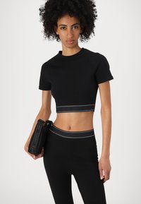 Fekete, crop szabású póló magas nyakkal és rövid ujjú, kontrasztos szalaggal, amely a márkanevet viseli. Magas derekú leggings-szel párosítva.