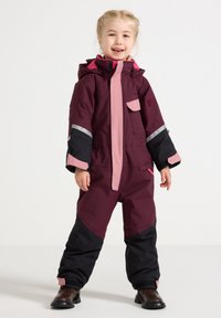 Bordeaux- en roze geïsoleerde sneeuwpak met zwarte accenten, voorzien van een capuchon, reflecterende strips en een voorzak. G gedragen met bruine laarzen.