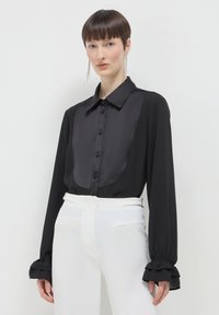 Blouse noire avec un panneau avant en satin, fermeture par boutons et poignets froncés, associée à un pantalon blanc taille haute. Texture lisse sur l'ensemble.