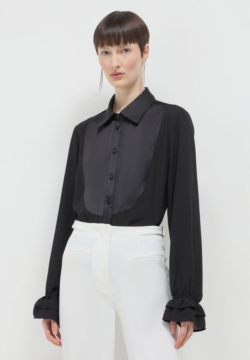 Schwarze Bluse mit einer Satin-Frontpartie, Knopfschluss und Rüschensäumen, kombiniert mit hochgeschnittenen weißen Hosen. Weiche Textur durchgehend.