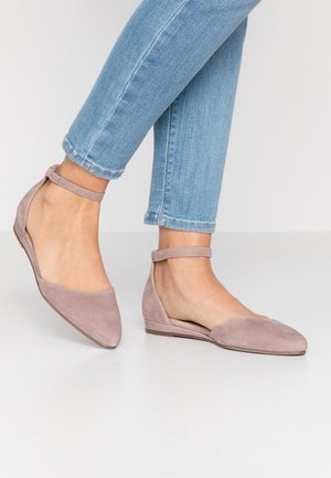 Füße einer Frau, die hell mauvefarbene spitz zulaufende Wildleder-Ballerinas mit Knöchelriemen trägt, kombiniert mit hellblauen Skinny-Jeans vor einem schlichten Hintergrund.