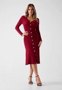 Robe en maille côtelée rouge avec manches longues, fermeture boutonnée sur le devant et fente à l'ourlet. Agrémentée de boutons dorés.