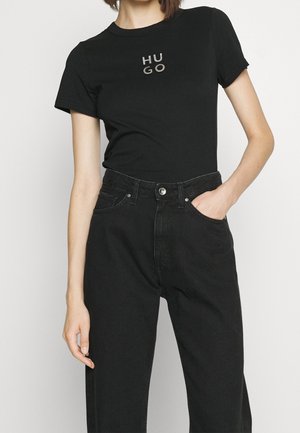 Kobieta ubrana w czarny T-shirt z krótkim rękawem z logo "HUGO" i czarne, wysokowystające dżinsy, jedną ręką w kieszeni na jednolitym tle.