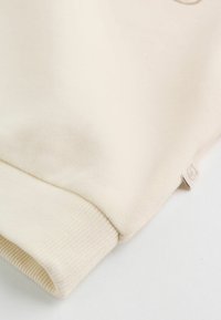 Sweter w kolorze kremowym, wykonany z materiału z prążkowanym dołem i mankietami rękawów. Mała haftowana metka na bocznej szwie. Miękka tekstura.