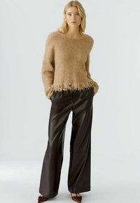 Beige Mohair-Pullover mit Fransen am Saum, kombiniert mit hochgeschnittenen, braunen Leggins aus Kunstleder mit weitem Bein, die Textur und Kontrast hinzufügen.