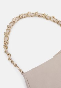 Beige kabelka s texturovaným povrchem a zlatým řetízkem na nošení, který obsahuje akcenty písmen. Řetěz je spojen s hladkými, lesklými detaily.