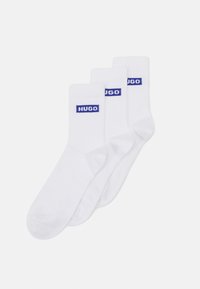 White cotton socks in a pack of three, featuring blue "HUGO" branding on the ribbed cuffs. Smooth texture and standard crew length.