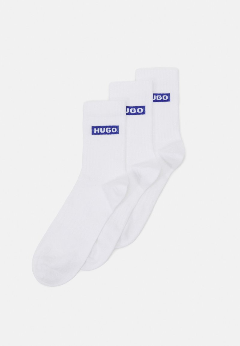 White cotton socks in a pack of three, featuring blue "HUGO" branding on the ribbed cuffs. Smooth texture and standard crew length.