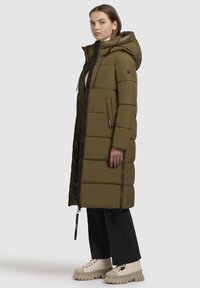 Olivovo zelený prešívaný dlhý kabát s kapucňou, ktorý má čierny zips a bočné vrecká. Nosený s čiernymi nohavicami a béžovými topánkami na robustnej podrážke.