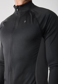 Chaqueta deportiva negra con cremallera hecha de tejido elástico. Cuenta con un cuello alto y un ajuste slim con costuras en contraste. Logotipo pequeño visible.