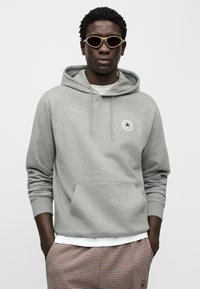 Γκρι φούτερ με κουκούλα και κορδόνια, τσέπη καγκουρό και στρογγυλό λογότυπο. Το μοντέλο φοράει μπεζ γυαλιά ηλίου και καρό παντελόνι με λευκό μανίκι.