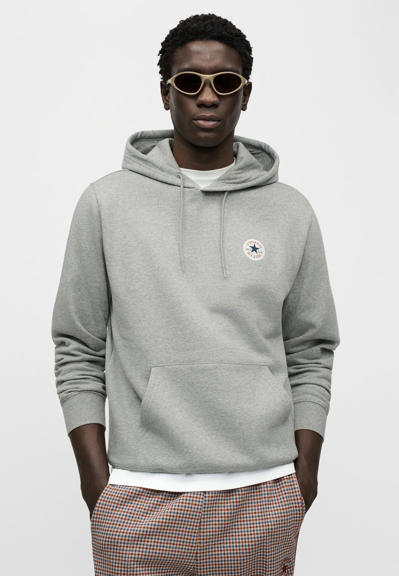 Grå hættetrøje med snore, kængurulomme og et rundt logo. Modellen bærer beige solbriller og ternede bukser med hvide manchetter.