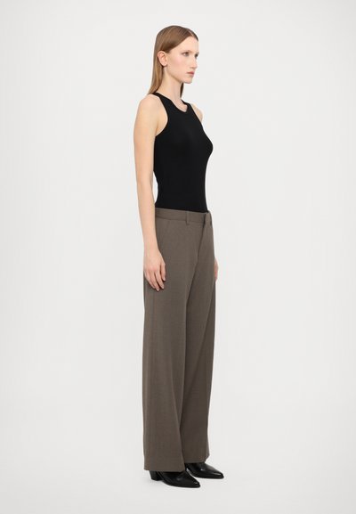 Body noir sans manches associé à un pantalon large taille haute marron. Le modèle porte des chaussures à talons noires. Arrière-plan simple et neutre.