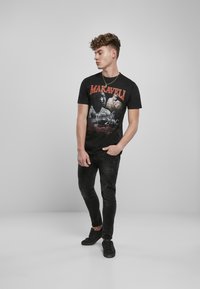 Czarna koszulka z grafiką z napisem "MAKAVELI" oraz wizerunkami związanymi z 2Paciem. Połączona z postrzępionymi czarnymi dżinsami i czarnymi sneakersami.