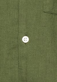 Tessuto di lino verde oliva con una trama sottile, caratterizzato da un bottone bianco con quattro fori, posizionato centralmente sul capo.