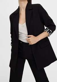 Czarna marynarka o dopasowanym kroju, zapinana na jeden guzik, z klapami z wycięciem. Posiada ozdobne mankiety z wzorem oraz boczne kieszenie.