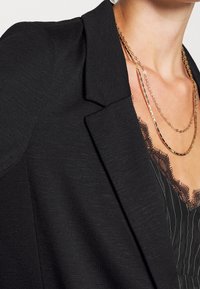 Czarna marynarka o fakturowanej tkaninie, z klapami i wyraźnie zarysowanymi ramionami. Widoczne paski warstwowych złotych naszyjników i koronkowe detale.