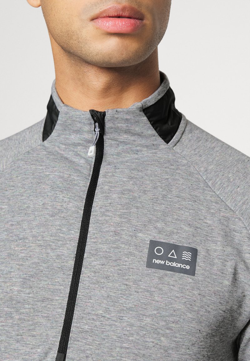 Chaqueta deportiva gris con cremallera y cuello alto, detalles en negro en el cuello, y un parche de logo con íconos y texto en el lado izquierdo del pecho.