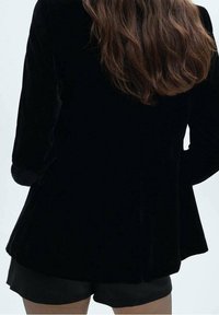 Blazer en velours noir à manches longues, doté d'un design évasé et d'une surface texturée. Vue de dos, associé à un bas noir court.