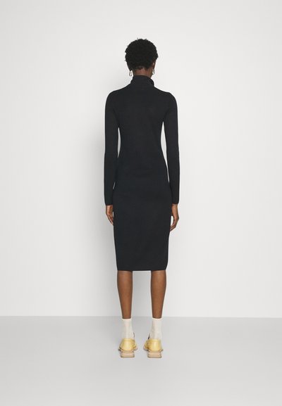 Μαύρο, μακρυμάνικο, φόρεμα με ψηλό λαιμό σε εφαρμοστή σχεδίαση, μέχρι το γόνατο με μαλακή υφή. Φορεμένο με κίτρινες παπούτσια και λευκές κάλτσες.