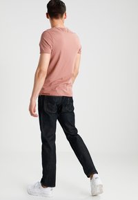 T-shirt di cotone rosa abbinato a jeans scuri e scarpe da ginnastica bianche, visto da dietro. La t-shirt ha una vestibilità classica e colletto a girocollo.