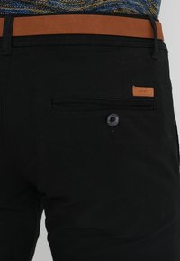 Svarta byxor med en bakficka som har knappstängning och en brun läderlogo. Midjebandet är brunt och texturerat.