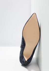Chaussure noire en daim à bout pointu montrant une semelle beige lisse avec un motif texturé antidérapant, positionnée verticalement contre des surfaces blanches.
