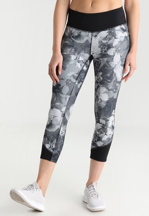 Kobieta ubrana w czarno-białe legginsy w kwiatowy wzór, sięgające powyżej kostek, oraz jasnoszare buty sportowe, stojąca z jedną stopą lekko wysuniętą do przodu.