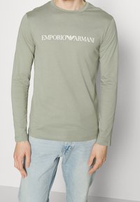 Mann trägt ein hellgrünes Langarmhemd mit weißem "EMPORIO ARMANI"-Text und Logo, kombiniert mit hellblauen Jeans.