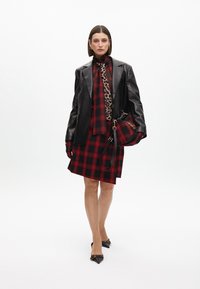 Čierny kožený blazer cez červené kárované šaty so zvieracím vzorom na kravate. Čierne špičaté podpätky a zodpovedajúca kabelka dokončujú outfit.