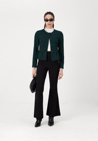 Green peplum jacket with buttons, white top, black flared pants, and ankle boots. Accessories include black handbag and slim sunglasses.