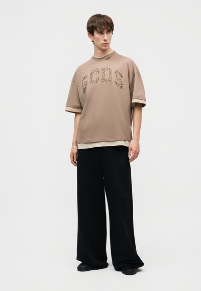 Νέος άνδρας που φοράει υπερμεγέθες μπεζ t-shirt με διάτρητο κείμενο και στρωμένο τελείωμα, πλατύ μαύρο παντελόνι και μαύρα παπούτσια, στέκεται σε λευκό φόντο.