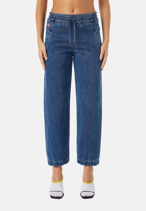 Kobieta nosząca wysokowodne niebieskie dżinsowe spodnie typu jogger z sznurkiem w talii i naszywanymi kieszeniami, zestawione z białymi sandałami na obcasie z żółtą lamówką.