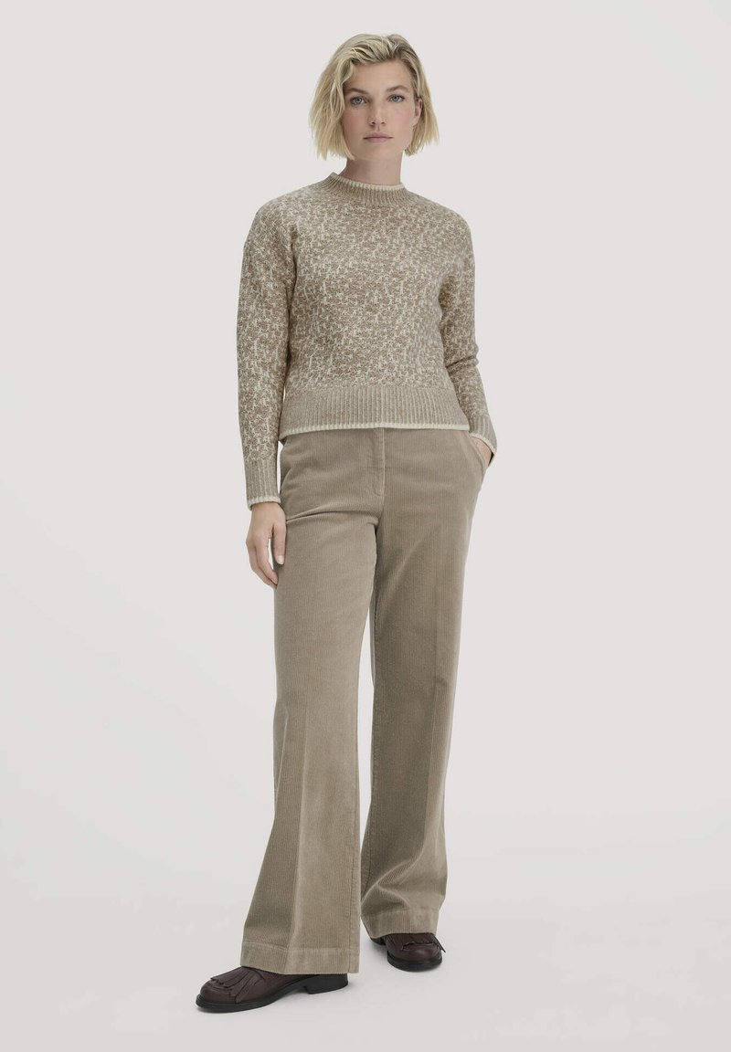 Beige gebreide trui met een textuurpatroon, gecombineerd met een wijde corduroy broek in een complementaire bruine tint. Het geheel is compleet met donkerkleurige schoenen.