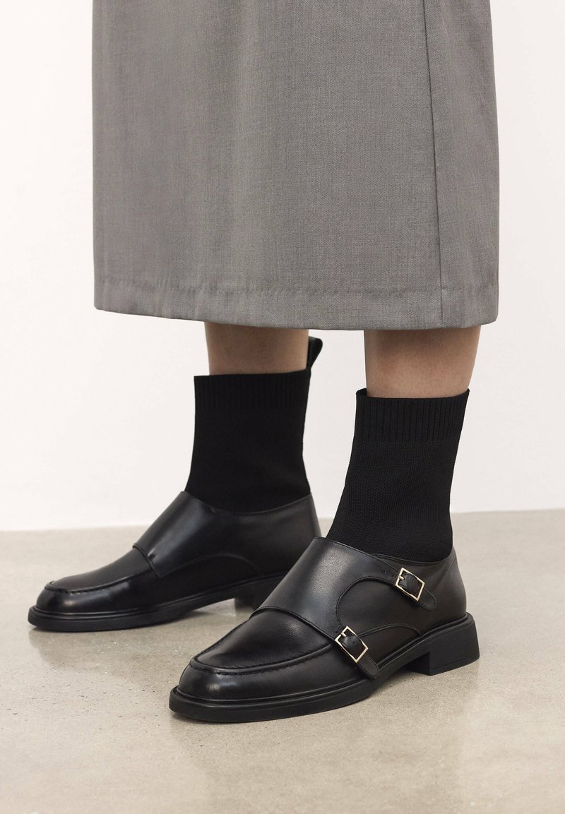 Schwarze Lederschuhe mit glatter Textur, ausgestattet mit zwei Schnallenakzenten, niedrigen Blockabsätzen und kombiniert mit schwarzen gerippten Socken.