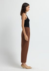 Jonge vrouw in profiel, gekleed in een zwart mouwloos topje, bruine hoge taillebroek en beige lage hakken, tegen een witte achtergrond.