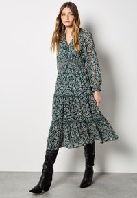 Largo vestido verde con un estampado de paisley, mangas largas, cintura fruncida, falda en capas y detalles de tul. Combinado con botas negras hasta la rodilla.