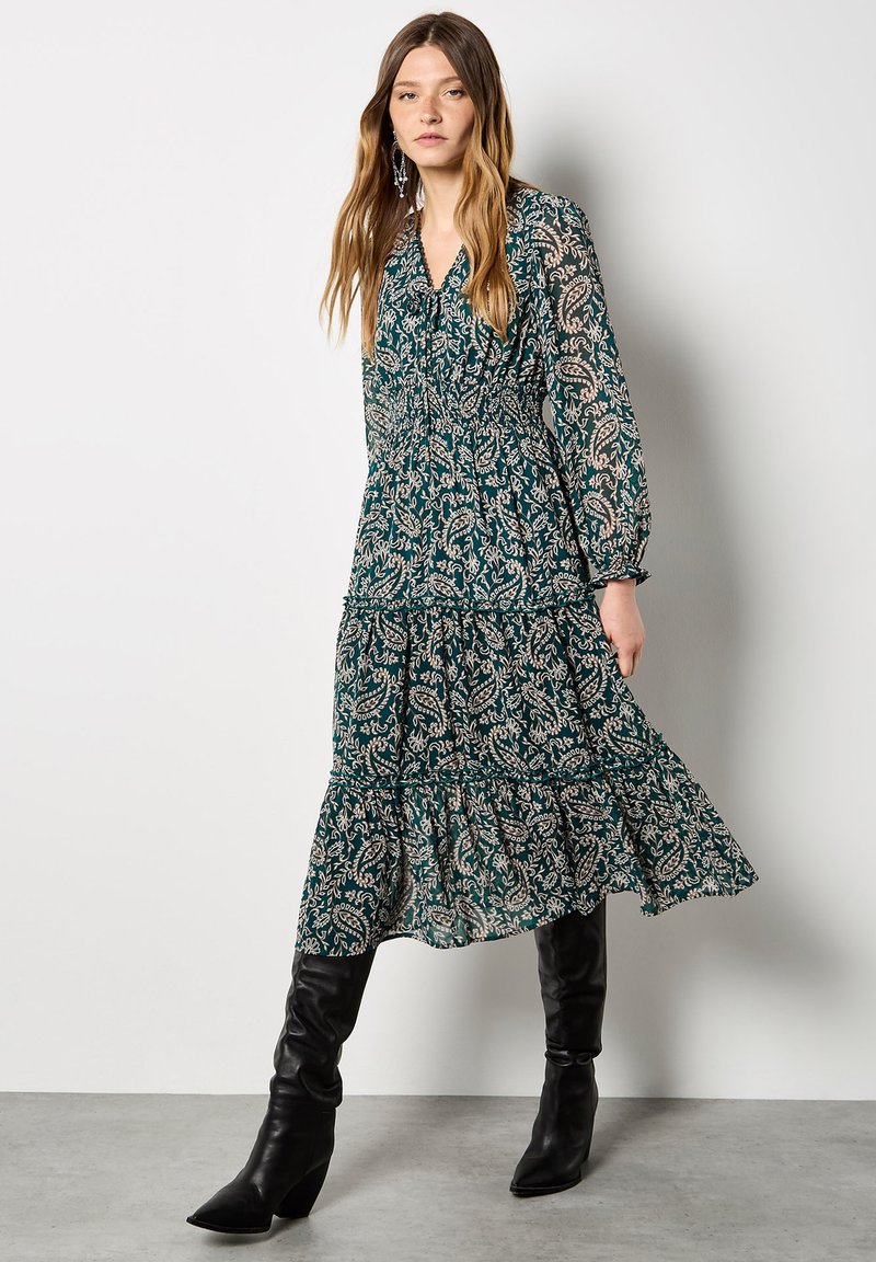 Largo vestido verde con un estampado de paisley, mangas largas, cintura fruncida, falda en capas y detalles de tul. Combinado con botas negras hasta la rodilla.