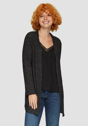 Cardigan noir, à fermeture ouverte, fabriqué en tissu doux et texturé. Superposé sur un haut noir avec une bordure en dentelle. Porté avec un jean bleu.
