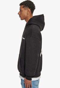Schwarzer Hoodie mit vorderer Kängurutasche, gerippten Bündchen und Saum, mit einem grafischen Logo auf der Rückseite und in einer lässigen Passform entworfen.