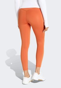 Oranžové športové legíny s hladkým povrchom, priliehavým strihom a kontrastnými švami, kombinované s bielymi teniskami na svetlom pozadí.