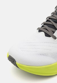 White athletic shoe with textured mesh upper, gray laces, and a bright yellow outsole. Features a rounded toe and streamlined design.