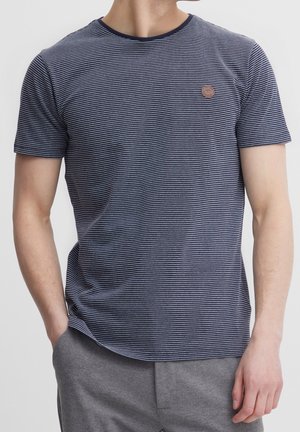 Mann mit navyblau-weiß dünn gestreiftem Kurzarm-T-Shirt und grauer Hose, mit der Hand in der Tasche, vor weißem Hintergrund.