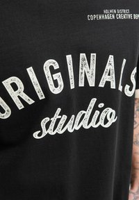 Camiseta negra con texto blanco que dice "ORIGINALS studio" y texto más pequeño "HOLMEN DISTRICT COPENHAGEN CREATIVE DEP" cerca del cuello.